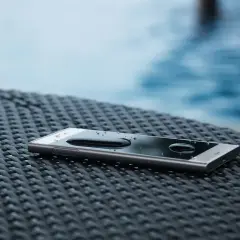 Smartfon z kroplami wody na ekranie, leżący na plecionej powierzchni. W tle basen. To pokazuje, co oznacza IP68 – odporność na wodę.