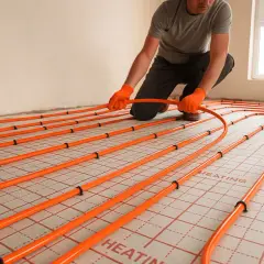Instalator układa pomarańczowe rury systemu ogrzewania podłogowego na specjalnych panelach.