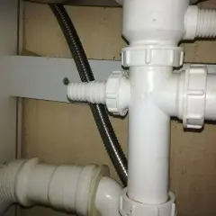 Biały plastikowy trójnik hydrauliczny pod zlewem, do którego podłączona jest elastyczna rura odpływowa i metalowy wąż.