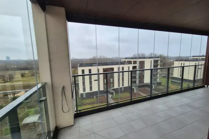 Zabudowa balkonu w bloku z widokiem na nowoczesne budynki. Szklane panele tworzą przestronną loggię.