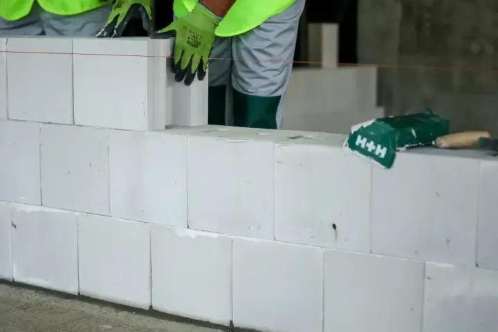 Budowa ściany z białych pustaków silikatowych. Ręce w zielonych rękawicach układają kolejny blok.