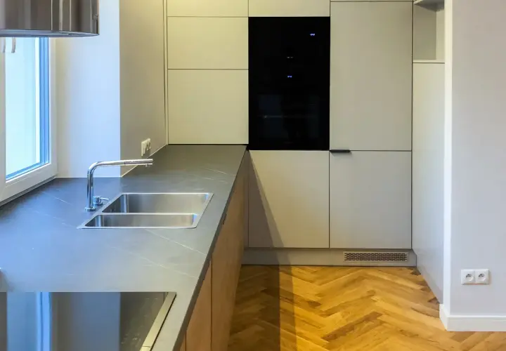 Nowoczesna kuchnia z szarymi frontami, wbudowanym piekarnikiem i podwójnym zlewozmywakiem. Podłoga w jodełkę i okap w kolorze RAL 9016 dodają elegancji.