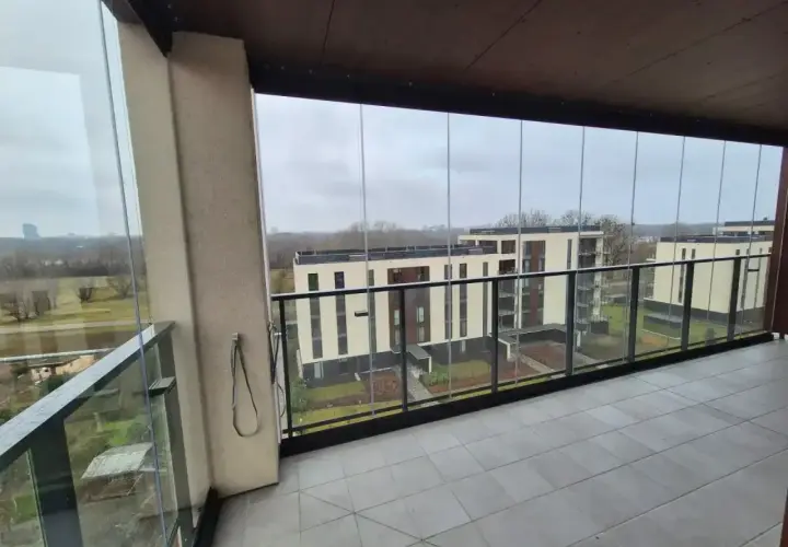 Zabudowa balkonu w bloku z widokiem na nowoczesne budynki. Szklane panele tworzą przestronną loggię.