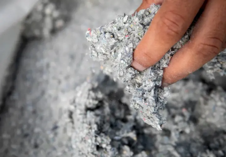Dłoń trzyma grudkę szarej, włóknistej masy. To materiał izolacyjny z recyklingu, oparty na celulozie.