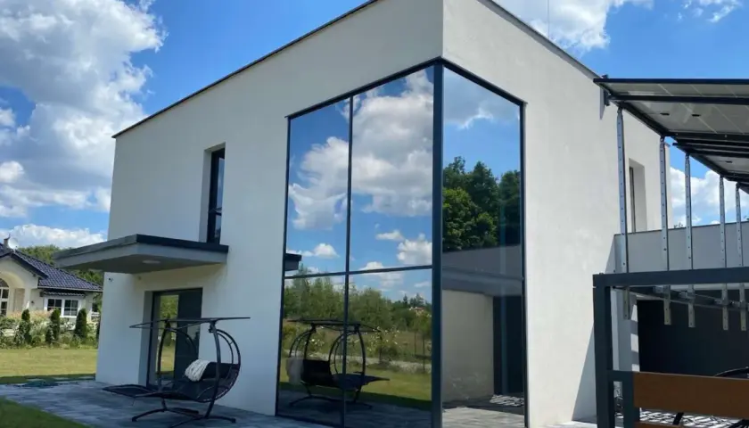 Nowoczesny dom z imponującym, narożnym oknem weneckim, w którym odbija się błękitne niebo z chmurami i zieleń drzew.