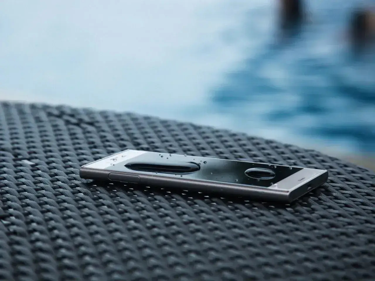 Smartfon z kroplami wody na ekranie, leżący na plecionej powierzchni. W tle basen. To pokazuje, co oznacza IP68 – odporność na wodę.
