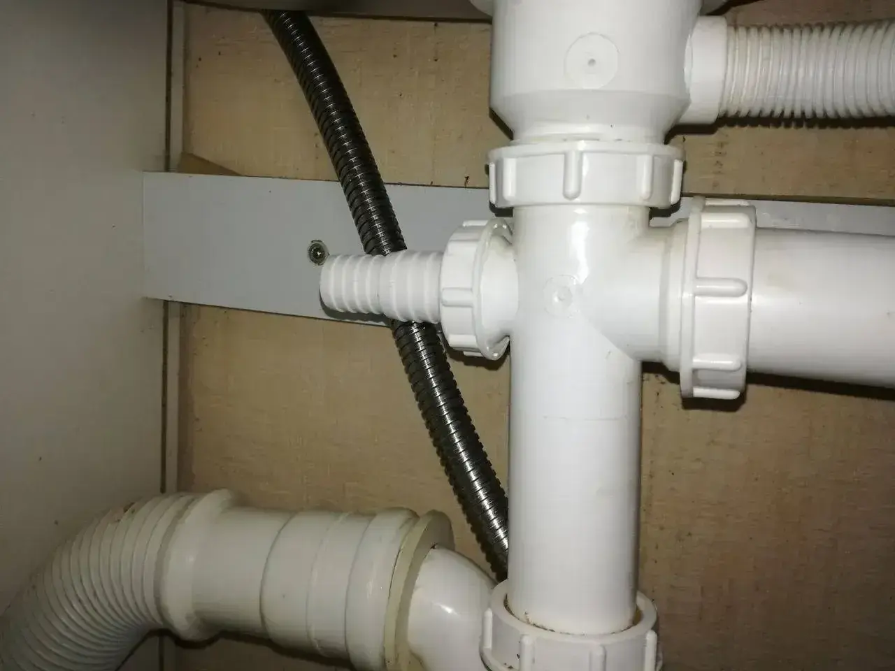 Biały plastikowy trójnik hydrauliczny pod zlewem, do którego podłączona jest elastyczna rura odpływowa i metalowy wąż.