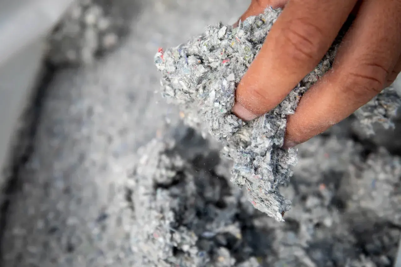 Dłoń trzyma grudkę szarej, włóknistej masy. To materiał izolacyjny z recyklingu, oparty na celulozie.