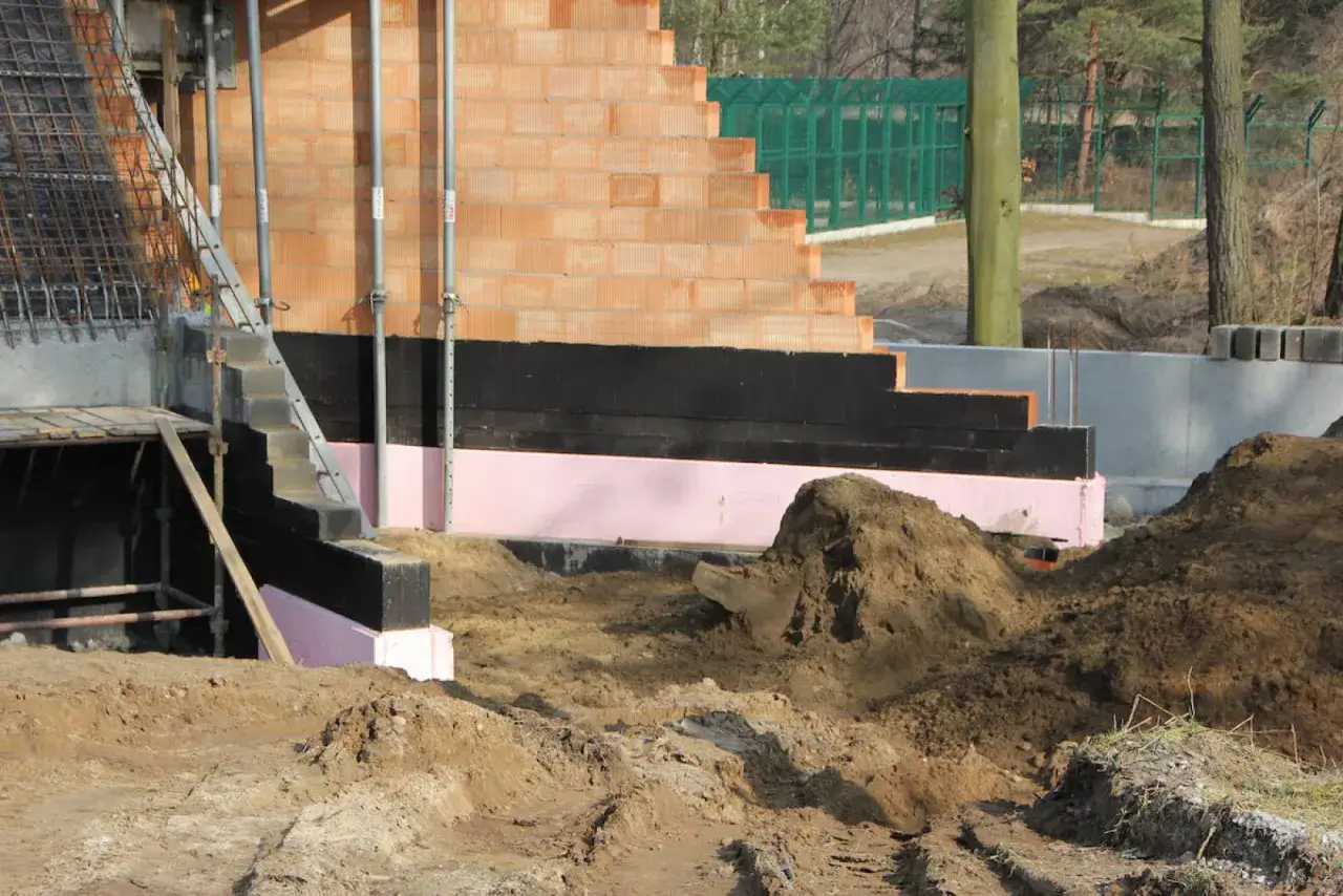 Budowa domu: różowy styropian EPS jako izolacja fundamentów, czarna hydroizolacja i ceglane ściany.